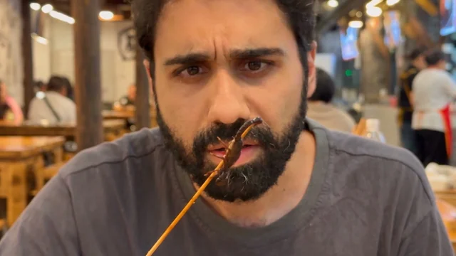 Beat 2: close-up, he studies a fried scorpion on a skewer, brow furrowed.
