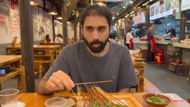 Beat 4: wider again, he chews and looks down at the tray, a bare skewer in his hand.