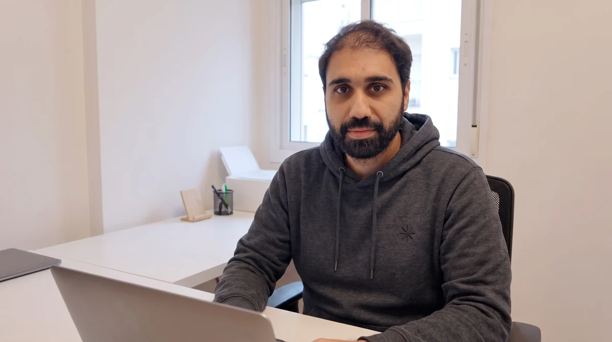 The wide look: the man in a dark grey hoodie at a white desk with a laptop, a bright window behind him.