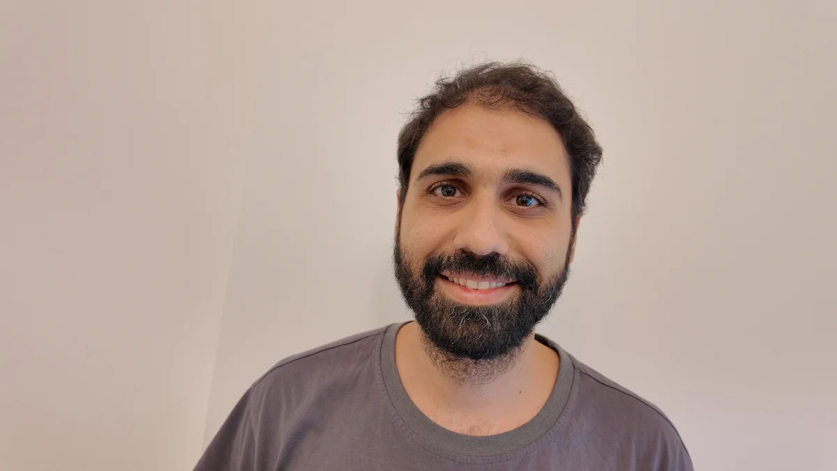 Front photo, smiling with his teeth showing, same shirt and wall.