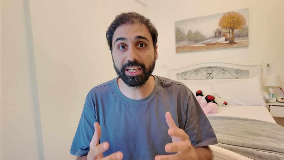 A real camera frame: the man in a blue t-shirt, sitting in front of a bed with a white scrolled metal headboard, plush toys on the pillows, a nightstand, and a painting of a tree on the wall.