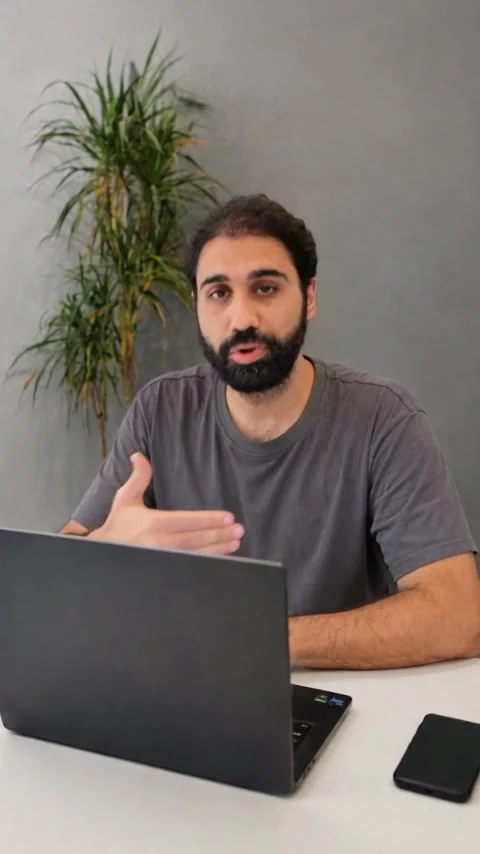 Vertical render: the man at a desk with a laptop, a plant behind him, framed close, his head and shoulders large in the frame.