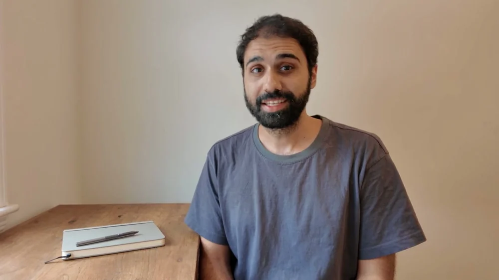 A Seedance render of the man in a grey t-shirt against a plain wall, smiling with his teeth showing, eyes on the camera.